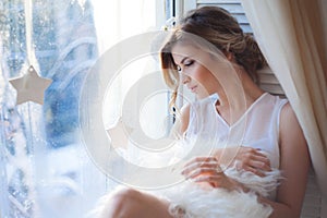 Young beautiful girl sitting on the windowsill, looking out window, morning light, glare