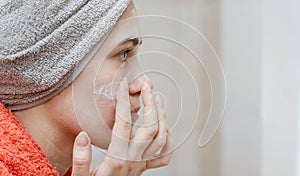 Young beautiful girl smearing her face with homemade natural cosmetic mask and looking in the mirror. Skin care and health care