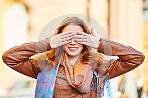 Young beautiful girl smiling and playing closes her eyes with her hands. Red-haired girl in a brown leather jacket on the street