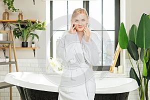 A young beautiful girl stands in the bathroom in a white robe touching her face in moisturizing patches