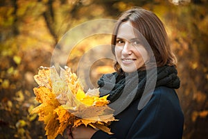 Young beautiful girl stands with a bouquet of maple orange leaves in the autumn park. They are looking at the camera and smiling.