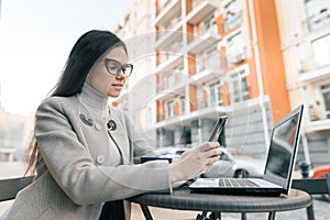 Young beautiful girl student with glasses in warm coat sitting in an outdoor cafe with laptop computer with phone cup of hot drink