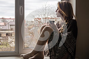 Young beautiful girl student sitting on a window sill at the window overlooking the city and drinking hot coffee from a mug