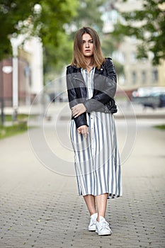 Young beautiful girl in stylish streetwear black leather jacket long striped dress white sneakers and with a fashionable bag strol