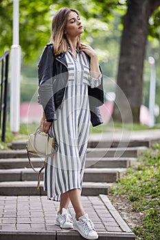 Young beautiful girl in stylish streetwear black leather jacket long striped dress white sneakers and with a fashionable bag strol