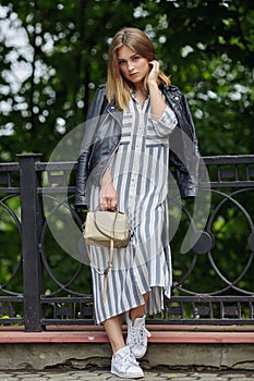 Young beautiful girl in stylish streetwear black leather jacket long striped dress white sneakers and with a fashionable bag strol