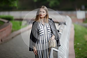 Young beautiful girl in stylish streetwear black leather jacket long striped dress white sneakers and with a fashionable bag strol