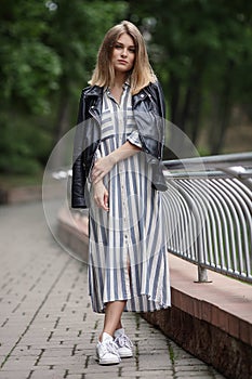 Young beautiful girl in stylish streetwear black leather jacket long striped dress white sneakers and with a fashionable bag strol