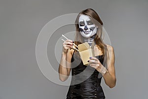 Young beautiful girl with a sugar skull make up, holds in her hand a paper box for instant noodles and chopsticks. Halloween face