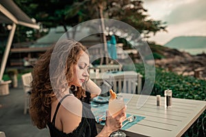 Young beautiful girl in summer bar with cocktail at sunset near the beach