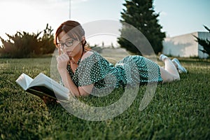 Young beautiful girl in the summer short dress reads a book lying on the grass.