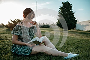 Young beautiful girl in the summer short dress reads a book sitting on the grass.