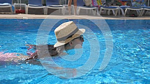 Young beautiful girl in sunglasses and hat swimming in pool. Woman relaxing in clear warm water on sunny day. Summer