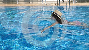 Young beautiful girl in sunglasses and hat swimming in pool. Woman relaxing in clear warm water on sunny day. Summer