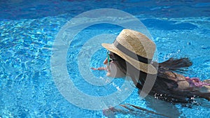 Young beautiful girl in sunglasses and hat swimming in pool. Woman relaxing in clear warm water on sunny day. Summer