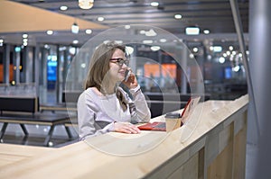 Young, beautiful girl talking on the phone standing at a table with a laptop and a cup of coffee at the airport