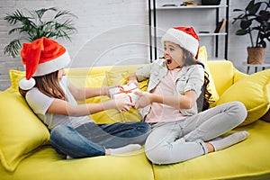 Young beautiful girl teenagers playing on yellow sofa. Together in room. Holding present box in four hands. Wear red hat