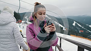 Young beautiful girl uses a mobile phone on the lift of a ski resort, mountains and skiers in the background.