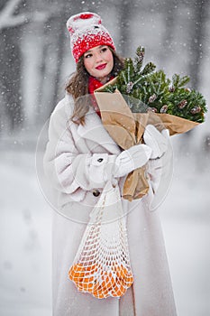 A young beautiful girl walks on a winter day in the garden in her hands holding tangerines and a bouquet of fir trees