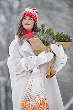 A young beautiful girl walks on a winter day in the garden in her hands holding tangerines and a bouquet of fir trees