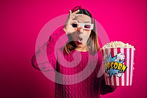 Young beautiful girl watching movie using 3d glasses eating box with popcorns doing ok gesture shocked with surprised face, eye