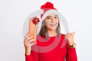 Young beautiful girl wearing Christmas Santa hat holding gift over isolated white background pointing and showing with thumb up to