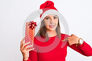 Young beautiful girl wearing Christmas Santa hat holding gift over isolated white background with surprise face pointing finger to