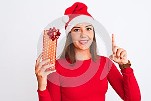 Young beautiful girl wearing Christmas Santa hat holding gift over isolated white background surprised with an idea or question