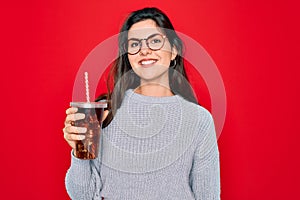 Young beautiful girl wearing glasses drinking sweet drink soda over red background with a happy face standing and smiling with a