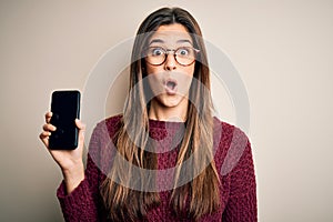 Young beautiful girl wearing glasses showing smartphone over isolated white background scared in shock with a surprise face,