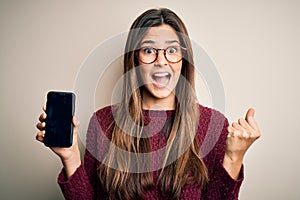 Young beautiful girl wearing glasses showing smartphone over isolated white background screaming proud and celebrating victory and