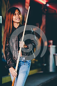 Young beautiful girl wearing leather jacket in a billiard club, with cue stick preparing for the game