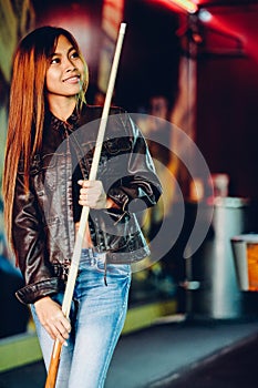 Young beautiful girl wearing leather jacket in a billiard club, with cue stick preparing for the game