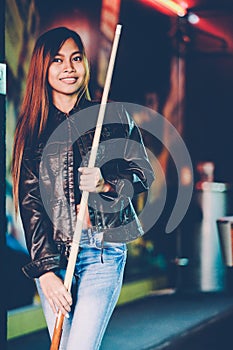 Young beautiful girl wearing leather jacket in a billiard club, with cue stick preparing for the game