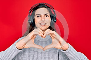 Young beautiful girl wearing modern headphones listening to music over red background smiling in love showing heart symbol and