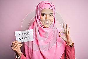Young beautiful girl wearing muslim hijab holding paper with fake news message doing ok sign with fingers, excellent symbol