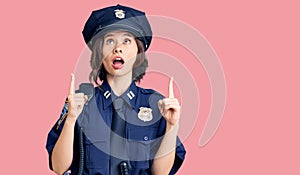 Young beautiful girl wearing police uniform amazed and surprised looking up and pointing with fingers and raised arms