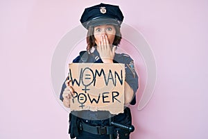 Young beautiful girl wearing police uniform holding woman power banner covering mouth with hand, shocked and afraid for mistake