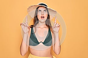 Young beautiful girl wearing swimwear bikini and summer sun hat over yellow background amazed and surprised looking up and