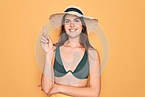 Young beautiful girl wearing swimwear bikini and summer sun hat over yellow background with a big smile on face, pointing with