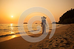 Young beautiful girl in a white dress and straw hat on a tropical beach at sunset. Summer vacation concept.