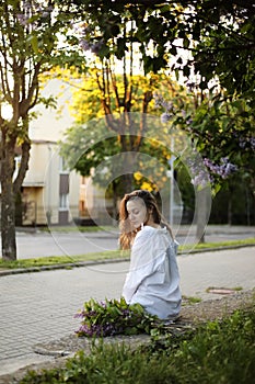 Young beautiful girl in a white shirt and black pant sitting near blooming lilac trees with purple flowers outdoors, flowering