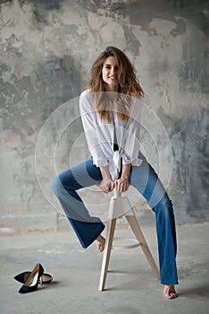 Young beautiful girl in a white shirt and flared jeans in a loft studio on a bar stool on a gray concrete background