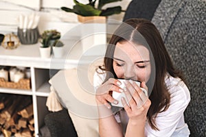 Young beautiful girl in white T-shirt and blue jeans drinks tea or coffee while sitting in chair at her cozy home.