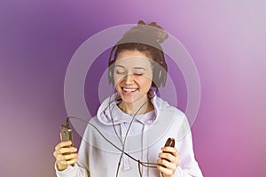 Young beautiful girl with white teeth listening to music on the phone wearing headphones in a sweatshirt on a purple