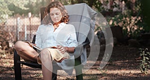 Young beautiful girl woman enjoys nature and reading a book in the summer in the garden of her house sitting on a chair