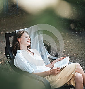 Young beautiful girl woman enjoys nature and reading a book in the summer in the garden of her house sitting on a chair