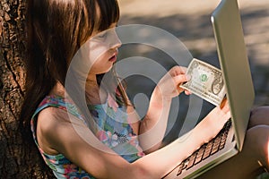 Young beautiful girl working with laptop and holding US Dollar bill as symbol of online payment and shopping. Summer vacation,