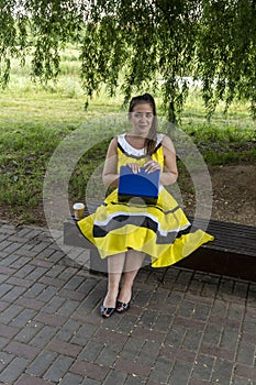 Young beautiful girl in a yellow dress with a laptop in the park. Works, learns, communicates online, making online shopping,