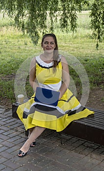 Young beautiful girl in a yellow dress with a laptop in the park. Works, learns, communicates online, making online shopping,